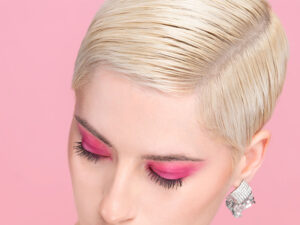 Primer plano de una persona de cabello rubio platino y corte corto con ojos cerrados maquillados en tonos fucsia, pestañas largas, pendientes brillantes y fondo rosa que transmite una apariencia profesional y segura.