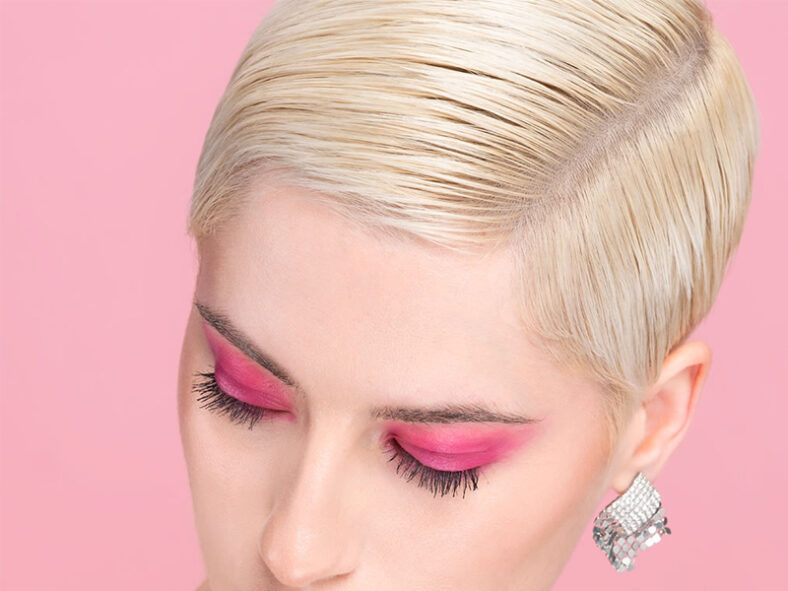 Primer plano de una persona de cabello rubio platino y corte corto con ojos cerrados maquillados en tonos fucsia, pestañas largas, pendientes brillantes y fondo rosa que transmite una apariencia profesional y segura.