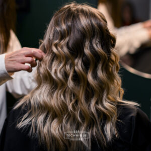 Cabello rubio con reflejos y ondas sueltas visto desde atrás mientras una mano de estilista lo arregla en un salón, mostrando un acabado elegante y profesional.