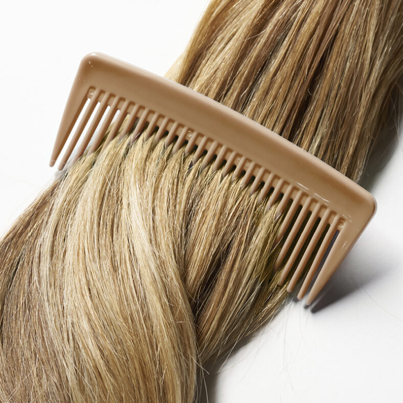 Peine beige deslizado por una mecha de cabello rubio liso sobre fondo blanco, detalle pensado para profesionales.