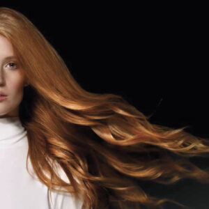 Mujer con rostro parcialmente visible y cabello rojo largo y ondulado que se extiende hacia la derecha sobre fondo negro, vistiendo cuello alto blanco, fotografía de alta calidad para presentación de producto.