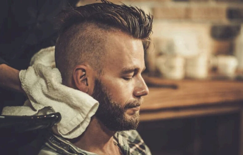 Hombre con corte degradado y barba cerrada, ojos cerrados mientras recibe atención en una barbería y le colocan una toalla blanca en la nuca, ambiente cálido y cuidado por profesionales.
