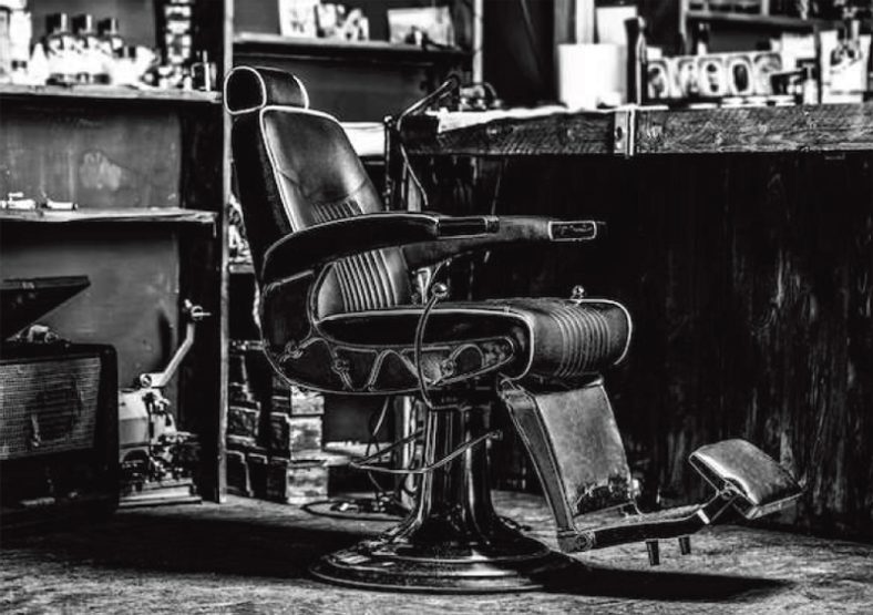 Silla de barbero vintage de cuero y metal con reposapiés reclinable en un interior de barbería con mostrador y estanterías, transmitiendo un entorno profesional.