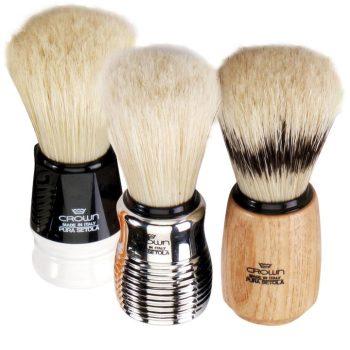 Tres brochas de afeitar con densas cerdas claras y mangos en plástico negro, metal cromado y madera natural, diseñadas para aplicación profesional en peluquería.