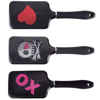 Tres cepillos de pala negros con mango ergonómico y cabezal rectangular forrado de cerdas y pedrería que forman un corazón rojo, una calavera con un corazón rosa en el ojo y las letras OX en rosa, accesorios llamativos para peluquería.