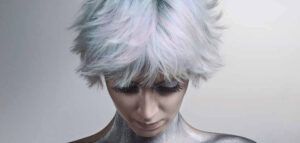 Persona con corte corto y esponjoso blanco plateado, mirada baja y pestañas largas, con cuello y hombros cubiertos de pintura corporal metálica en una imagen de estudio profesional.