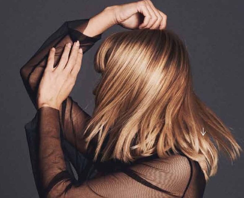 Persona de perfil con cabello rubio hasta los hombros en movimiento y manos elevadas, vistiendo una blusa negra transparente sobre fondo gris, fotografía editorial que destaca un corte y color profesional y un acabado completo.