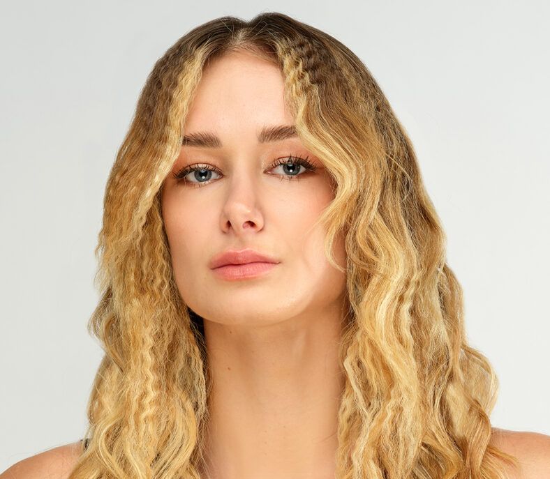 Mujer de rasgos suaves y piel clara con cabello rubio largo texturizado en ondas y crimpados, mirada serena y maquillaje natural sobre fondo neutro, ideal para catálogo de peluquería.