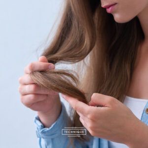 Mujer revisando las puntas de su cabello largo y castaño con las manos que muestran textura y posibles puntas abiertas, en una imagen de alta calidad con iluminación suave y apariencia de tratamiento autorizado.