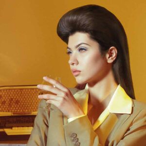 Mujer joven con peinado retro y maquillaje azul, vestida con traje beige y solapa amarilla satinada, sostiene una copa de cristal y mira de lado frente a un fondo mostaza con una radio vintage, transmitiendo un aire 'profesional'.