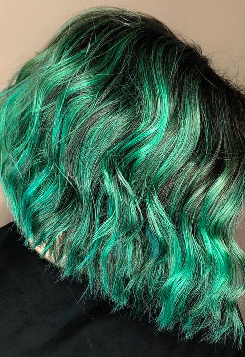 Cabello corto ondulado visto de perfil con raíces oscuras y reflejos verde esmeralda vibrantes en contraste sobre fondo neutro, fotografía de 'alta calidad'.