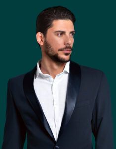 Retrato de un hombre de barba y cabello oscuro con traje de solapas satinadas, camisa blanca y mirada lateral sobre fondo verde azulado, con aspecto profesional.