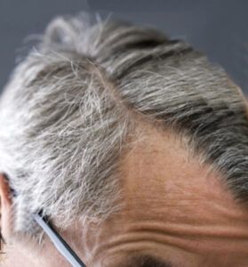 Primer plano de cabello canoso con raya lateral y frente arrugada junto a la patilla de unas gafas, que sugiere peluquería y evoca la presencia de una herramienta de estilismo.