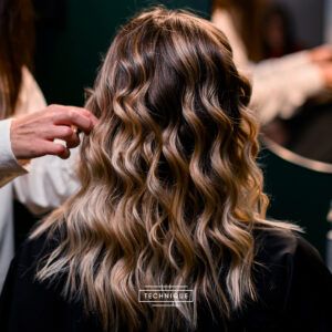 Cabello ondulado con mechas rubias visto desde atrás mientras una mano de estilista lo acomoda en un salón, mostrando un acabado profesional y sutilmente orientado a la venta.