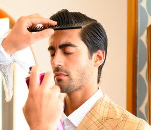 Hombre con los ojos cerrados recibe un peinado mientras una mano lo peina con un peine y otra aplica spray en una peluquería, vistiendo camisa blanca y chaqueta a cuadros.