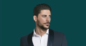Hombre adulto joven con cabello oscuro peinado hacia atrás y barba recortada, vistiendo camisa blanca y chaqueta oscura sobre fondo verde azulado, imagen pulida orientada a peluquería.