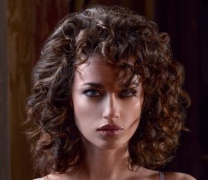 Primer plano de una mujer de rasgos definidos con cabello castaño rizado, mirada fija y expresión seria, maquillaje sutil y iluminación lateral que realza el rostro en un retrato profesional.