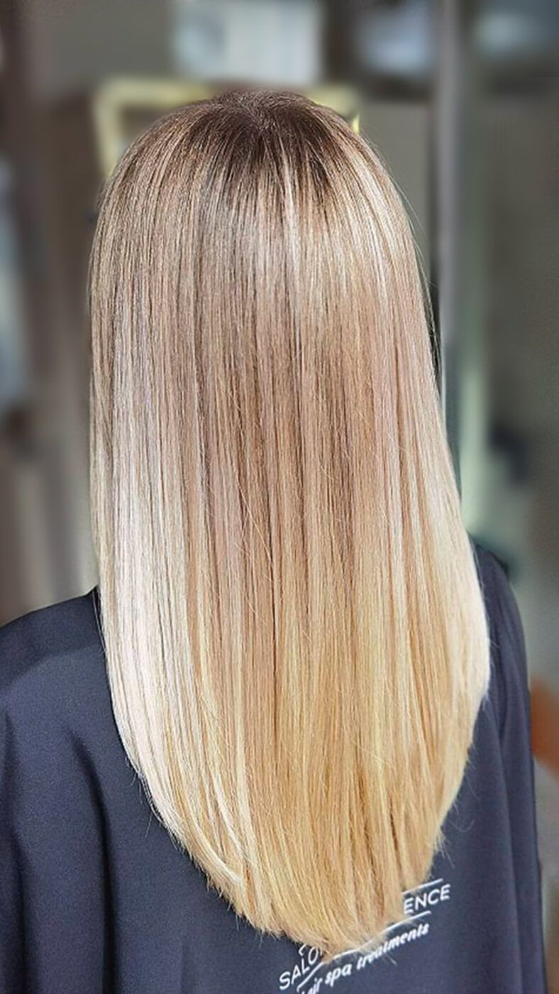 Vista posterior de cabello largo, liso y rubio con raíces algo más oscuras, corte recto y brillo uniforme sobre una capa negra en un entorno desenfocado de peluquería, gratis.