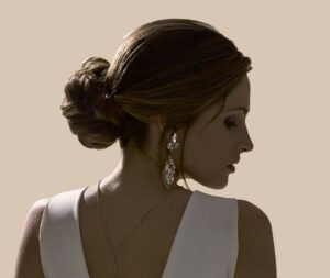 Mujer de perfil con recogido bajo y vestido blanco sin mangas, pendientes largos y collar delicado sobre piel iluminada contra un fondo neutro, de aspecto elegante y profesional, disponible para catálogo.
