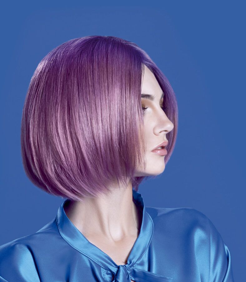Mujer en perfil con corte bob liso color púrpura, piel clara y maquillaje suave, vistiendo una blusa satinada azul sobre fondo azul, fotografía estilizada y 'profesional'.