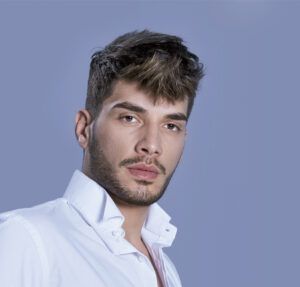 Retrato en primer plano de un hombre joven de rasgos definidos con barba corta, cabello castaño peinado hacia adelante y mirada neutra, vistiendo una camisa blanca de cuello abierto sobre un fondo liso azul grisáceo, imagen de 'alta calidad'.