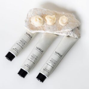 Presentación minimalista de tres tubos plateados de producto cosmético con tapas negras, apoyados por dos piedras claras coronadas por tres montículos de crema beige, imagen de 'alta calidad'.
