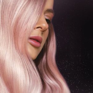 Primer plano de una persona con cabello largo y liso rosa pastel que cae sobre un costado del rostro, piel impecable y labios rosados brillantes sobre un fondo oscuro con pequeños destellos, imagen de alta calidad.