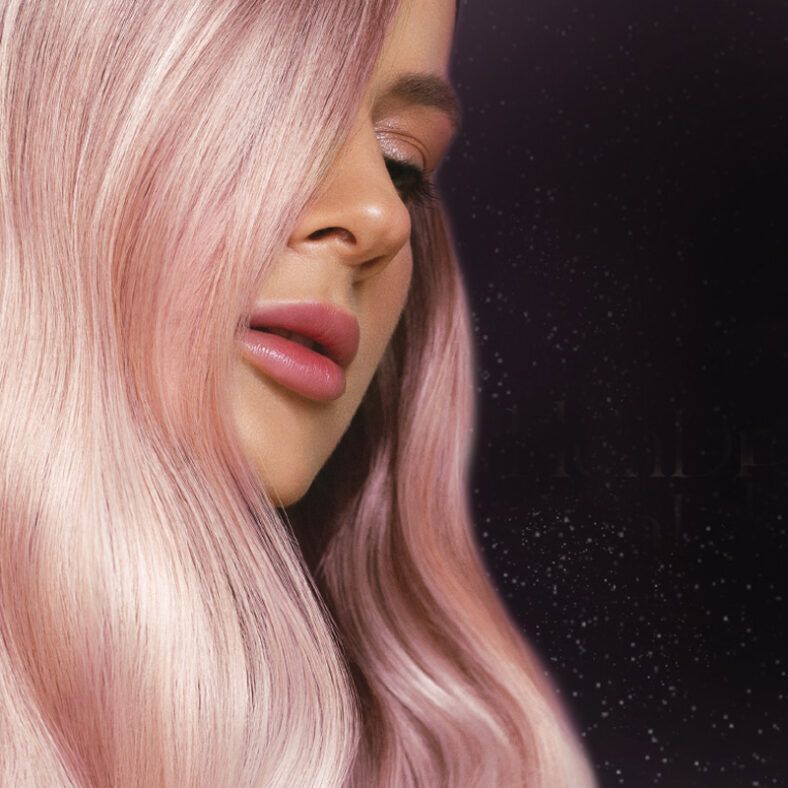 Primer plano de una persona con cabello largo y liso rosa pastel que cae sobre un costado del rostro, piel impecable y labios rosados brillantes sobre un fondo oscuro con pequeños destellos, imagen de alta calidad.
