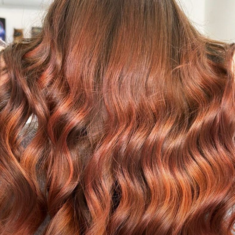 Cabello largo y ondulado en ricos tonos cobre y caoba con reflejos cálidos y brillo sedoso, acabado de peluquería.