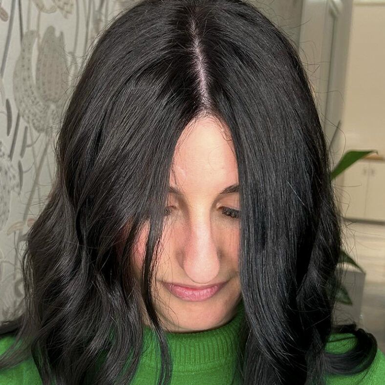 Persona de cabello largo y oscuro con raya al medio mirando hacia abajo y con suéter verde en un interior, retrato de alta calidad.