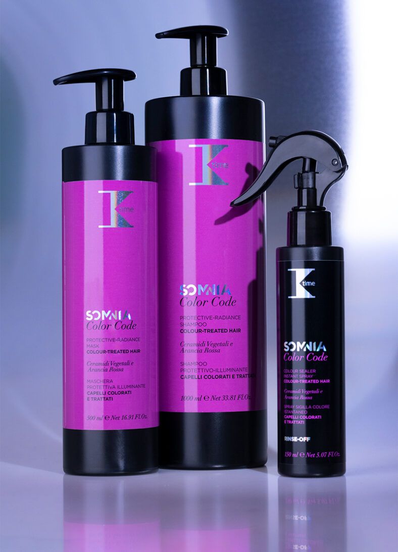 Tres envases de productos para cabello coloreado dispuestos en estudio: dos botellas altas con dosificador y etiqueta magenta junto a un frasco pulverizador negro más pequeño, con acabado brillante que sugiere uso profesional.