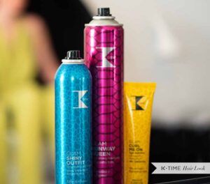 Tres envases de productos capilares con acabado metálico y estampado —un spray azul, un spray rosa alto y un tubo amarillo— en primer plano sobre un fondo de salón desenfocado, transmitiendo un uso profesional.