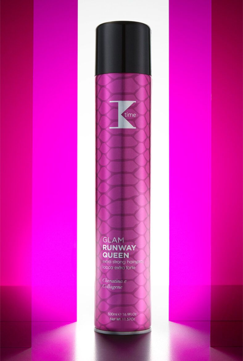 Lata aerosol alta color fucsia metalizado con tapa negra y patrón tipo escamas, etiquetada "Glam Runway Queen" como laca fijadora extra fuerte sobre fondo iluminado en tonos rosa, mejor precio.