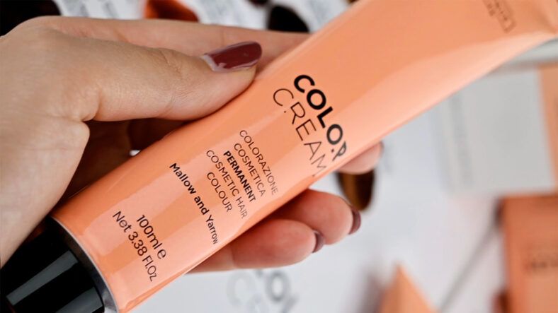 Mano con uñas barnizadas sostiene un tubo salmón de crema colorante para cabello con texto impreso en un primer plano nítido que transmite alta calidad.