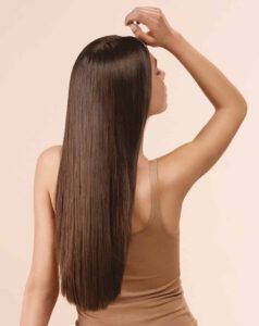 Mujer vista de espaldas con cabello largo, liso y brillante de color castaño, vistiendo una camiseta sin mangas beige y levantando un brazo sobre un fondo neutro, imagen ideal para promoción de peluquería.