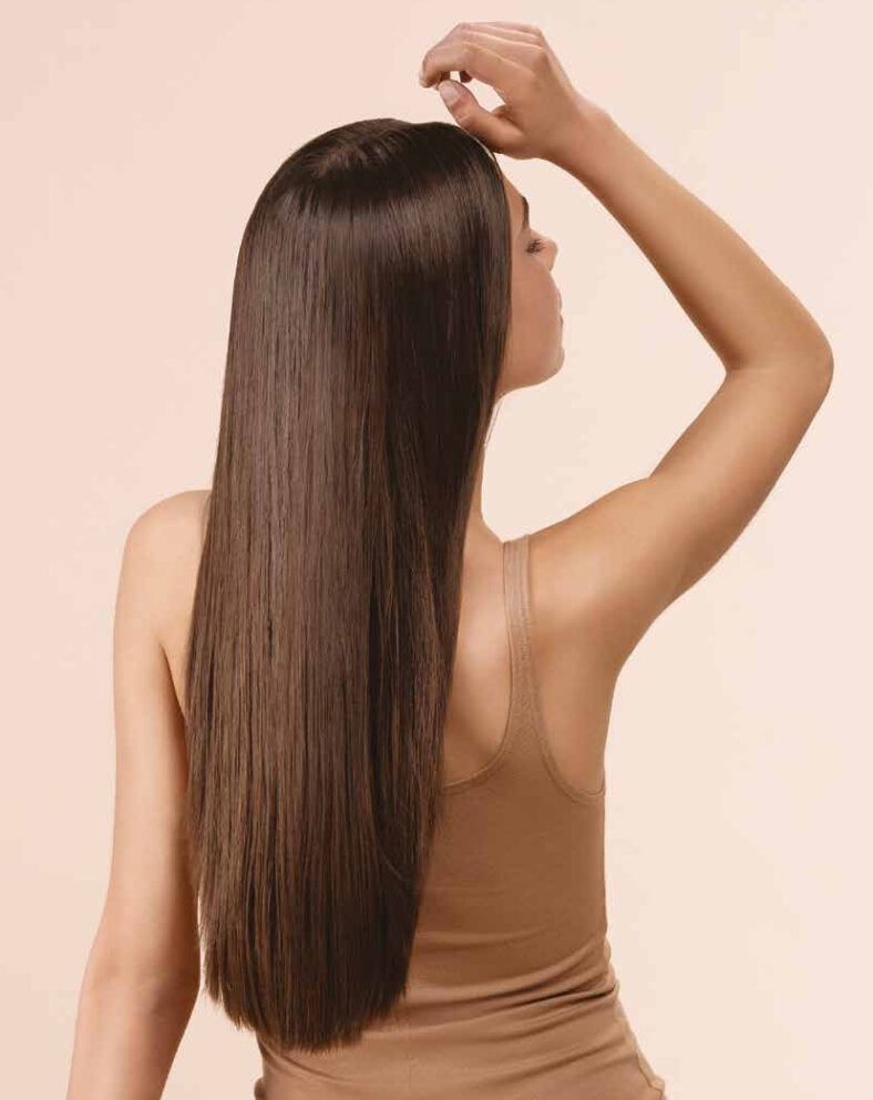 Mujer vista de espaldas con cabello largo, liso y brillante de color castaño, vistiendo una camiseta sin mangas beige y levantando un brazo sobre un fondo neutro, imagen ideal para promoción de peluquería.