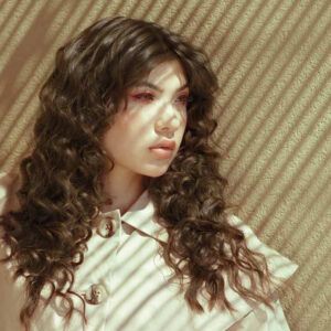Persona de cabello castaño rizado con maquillaje suave mirando hacia la derecha, vestida con abrigo color marfil con botones grandes, iluminada por franjas de luz que crean sombras diagonales sobre su rostro y un fondo texturizado, imagen de alta calidad.