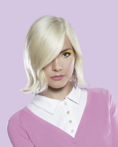 Mujer con cabello rubio platino corto y flequillo lateral, maquillaje con sombra amarilla y labios rosados, pendientes largos y camisa blanca bajo un cárdigan rosa sobre fondo lila, fotografía de alta calidad que ilustra un producto de moda.