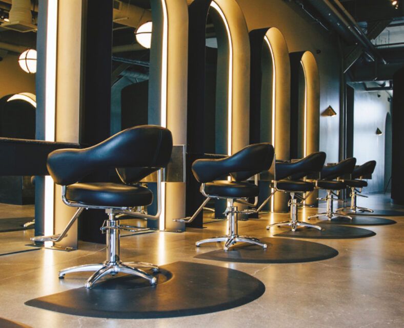 Sillas de peluquería negras alineadas frente a espejos arqueados retroiluminados en un salón moderno con acabados cromados y suelo pulido, que transmiten alta calidad y un diseño único.