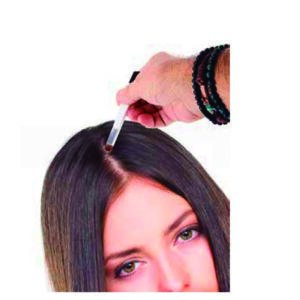 Mano con pulseras sostiene un aplicador junto a la raya del cabello de una mujer de pelo lacio y oscuro durante la aplicación de un tratamiento capilar, imagen pensada para uso profesional y mayorista.