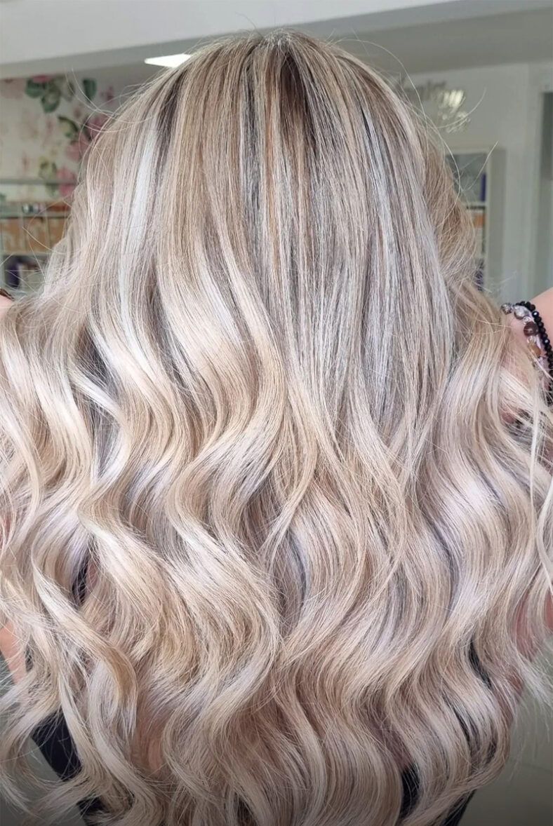 Cabello largo y ondulado con mechas rubias frías y cálidas tipo balayage, acabado brillante y movimiento, mostrado desde la nuca en una peluquería.